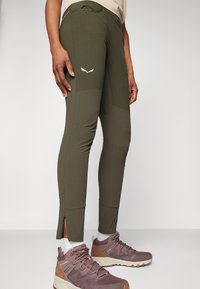 Olive green performance pants with a textured design and side zippers at the ankles. Features a white logo detailing on the thigh.