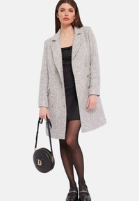 Cappotto grigio in tweed doppiopetto con bottoni dorati, indossato sopra un vestito nero. Include una borsa a tracolla nera con logo e collant neri.