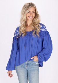 Blusa blu fluida con scollatura e maniche a volants, decorata con ricami floreali sulle spalle. Abbinata a jeans blu chiaro.
