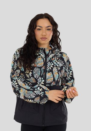 Kobieta poprawiająca rękaw czarnej kurtki z kolorowym, abstrakcyjnym nadrukiem liści i zwierząt, stojąca na tle jednolitego jasnoszarego tła.