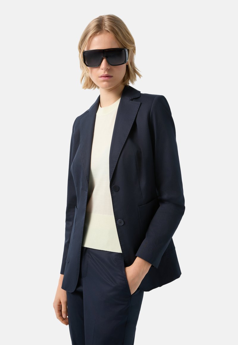 Námořnický blazer se dvěma knoflíky, spárovaný s bledým vrškem a odpovídajícími kalhotami. Velké černé sluneční brýle dotvářejí celkový vzhled.