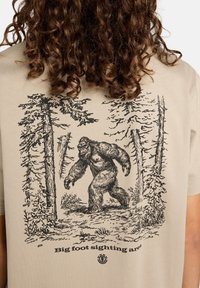 Camiseta beige con una ilustración negra de un hombre de las nieves en un bosque, con árboles altos y el texto "Área de avistamiento de Bigfoot" debajo.