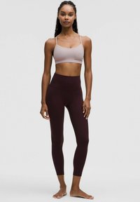 Lichtroze sportbeha met dunne bandjes, gedragen met hooggetailleerde bordeauxrode leggings. Het materiaal lijkt glad en aansluitend.