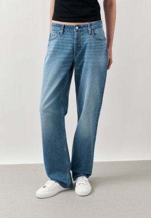 Person iført lyseblå jeans med vide ben og hvite sneakers, står foran en ensfarget lys grå bakgrunn.