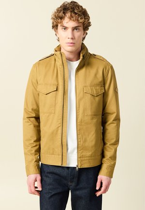 Joven con cabello rizado que lleva una chaqueta mostaza sobre una camisa blanca y jeans azul oscuro, posando sobre un fondo beige liso.