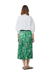 Witte blouse met lange mouwen en hoge hals, gecombineerd met een groene plissérok met bloempatroon en zwarte flats.
