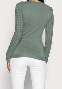 Femme portant un pull ajusté vert sauge avec des manches texturées et un pantalon blanc, se tenant de dos face à l'objectif sur un fond uni.