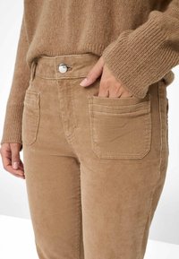 Nærbillede af en person iført beige corduroy bukser med rektangulære for- lommer og en matchende striktrøje, hvor hånden delvist er i lommen.