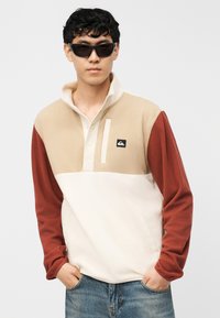 Νέος άνδρας που φοράει μαύρα γυαλιά ηλίου, μια μπλούζα φλις σε μπεζ, κρεμ και σκουριά χρώματα και μπλε τζιν, στέκεται με τα χέρια στις τσέπες.