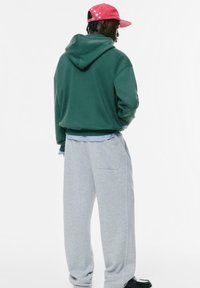Felpa verde con tasca frontale, abbinata a pantaloni grigi. Il modello indossa un cappellino rosso con un motivo di stelle bianche.