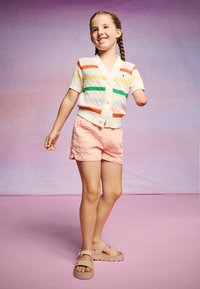 Stickad cardigan med flerfärgade ränder, krämfärgad bas, tillsammans med ljusrosa texturerade shorts och beiga sandaler, stående framför en rosa bakgrund.