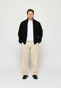 Ein Mann steht mit den Händen in den Taschen, trägt eine schwarze Jacke, ein weißes Hemd, beige Hosen und schwarze Schuhe vor einem schlichten hellen Hintergrund.