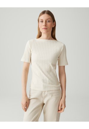 Femme aux cheveux lisses châtain clair portant un haut blanc à manches courtes côtelé et un pantalon beige clair, debout devant un fond neutre.