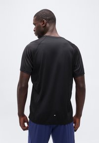 Άνδρας που φοράει μαύρο αθλητικό μπλουζάκι με κοντά μανίκια και μπλε σορτς, γυρισμένος πλάτη, σε απλό λευκό φόντο.