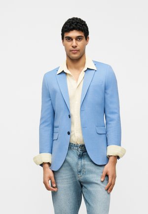 Homme portant un blazer bleu clair sur une chemise jaune pâle et un jean bleu clair, debout devant un fond blanc uni.