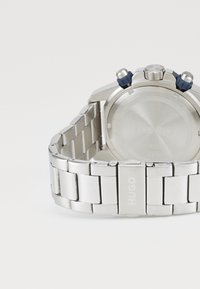 Stainless steel watch with a silver bracelet, featuring a round bezel, blue accents, and a polished finish. Includes the brand name "Hugo" engraved.