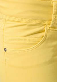 Žluté džíny mají hladkou látku se šitými kapsami, stříbrným knoflíkem a těsným střihem. Materiál je měkký a elastický.