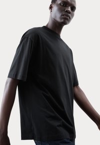 Maglietta nera in cotone leggero con maniche corte, vestibilità ampia e colletto rotondo classico. Design minimalista senza motivi o dettagli visibili.