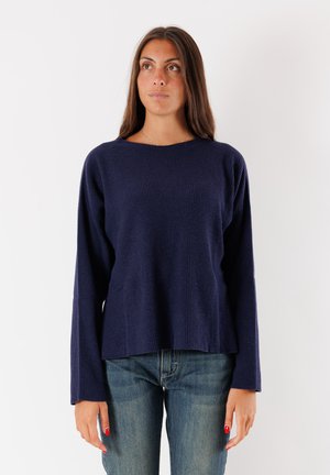 Donna con lunghi capelli castani che indossa un maglione a maniche lunghe blu navy a coste e jeans blu, in piedi di fronte a uno sfondo bianco semplice.