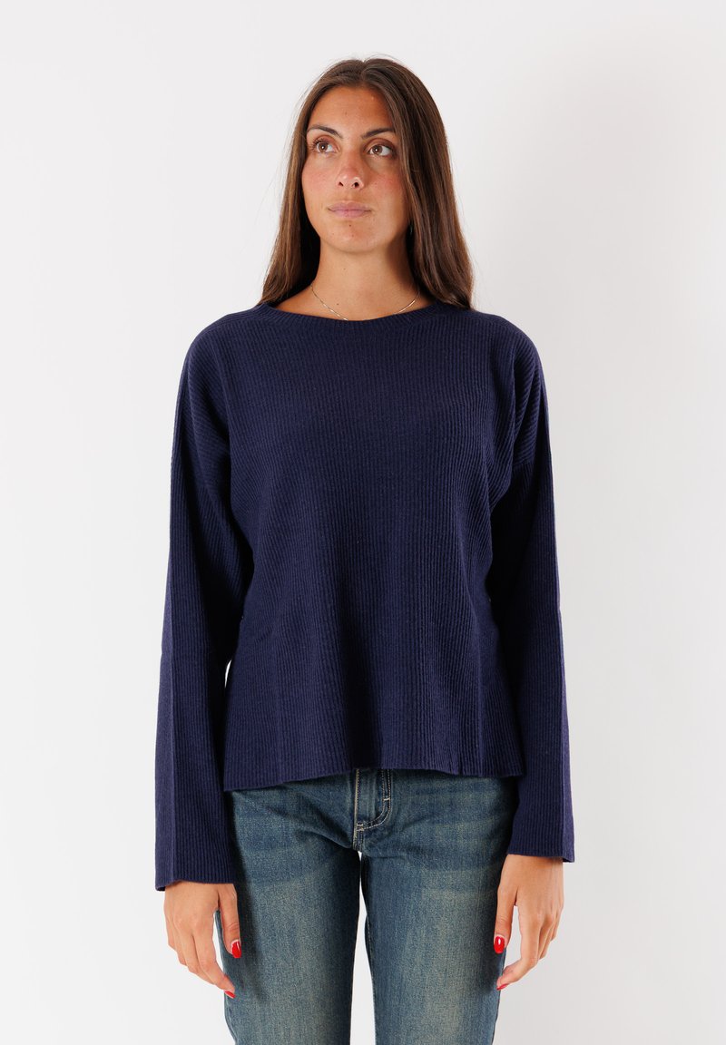 Donna con lunghi capelli castani che indossa un maglione a maniche lunghe blu navy a coste e jeans blu, in piedi di fronte a uno sfondo bianco semplice.