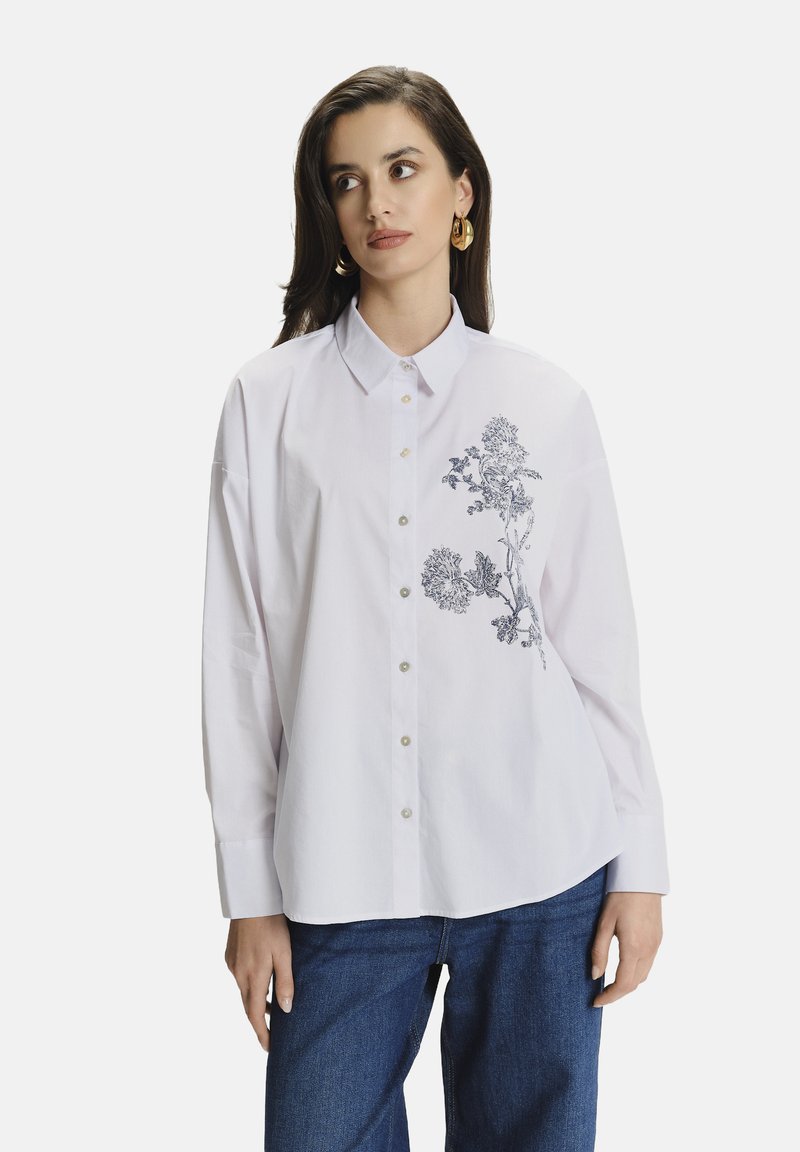 Camisa blanca de manga larga con cuello abotonado, que presenta bordados florales azules en el lado izquierdo y un corte relajado.