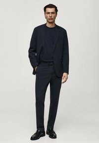 Abito scuro su misura con una camicia aderente coordinata. Il materiale appare liscio e raffinato. I dettagli di accentuazione includono una tasca sulla giacca.