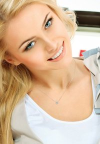 Collier en argent avec un petit pendentif, chaîne délicate, porté par une personne aux longs cheveux blonds. Chemise blanche unie, éclairage naturel doux.