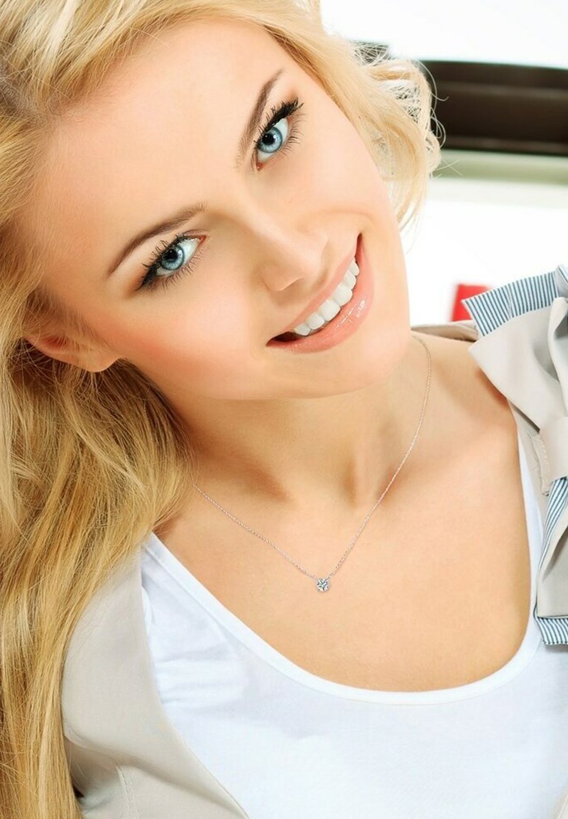 Collier en argent avec un petit pendentif, chaîne délicate, porté par une personne aux longs cheveux blonds. Chemise blanche unie, éclairage naturel doux.