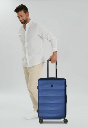 Mann in weißem Hemd und beiger Hose zieht einen blauen Hartschalenkoffer mit vier Rollen vor weißem Hintergrund.