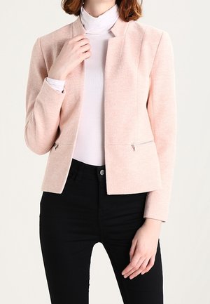 Femme portant un blazer rose clair avec des poches zippées sur un col roulé blanc et un pantalon noir, posant devant un fond uni.