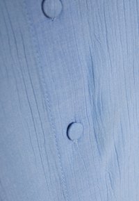 Ljusblå skjorttyg med en texturerad, krinklad yta. Har två matchande knappar och tonade sömmar längs knappslån.