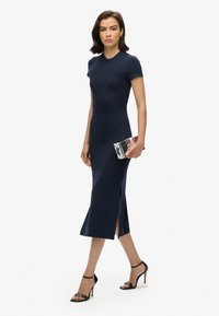 Robe midi navy à manches courtes, corsage ajusté, fentes latérales et texture lisse. Associée à des escarpins noirs à lanières et à une pochette argentée.