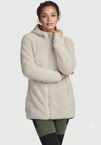 Huppari, jossa on fleece-kangas ja vaalea beige väri, vetoketjukiinnitys, pehmeä pinta ja sivutaskut, yhdistettynä tumman vihreisiin legginsseihin.