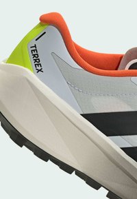 Chaussure de sport grise et blanche avec des rayures noires, des accents orange et un talon vert vif. Caractérisée par un tissu texturé et une semelle profilée.