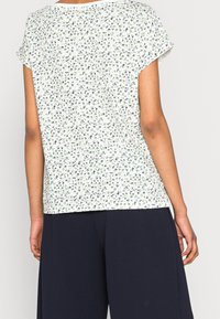 Vrouw in een kortoeven witte blouse met een klein groen en marineblauw bloemenpatroon, gecombineerd met losse, donker marineblauwe pantalon, achterkantzicht.