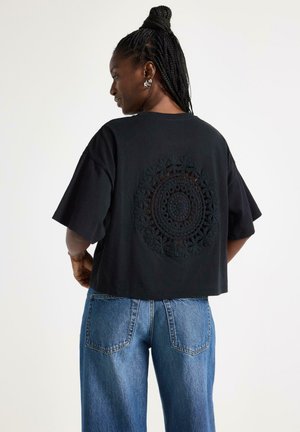 Kvinde med flettet hår iført store øreringe, en sort oversized T-shirt med et rundt hæklede mønster på bagsiden og blå jeans.