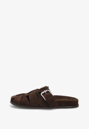 Sandalia marrón de ante sin cordones con tiras anchas y una hebilla plateada en el lateral, suela plana y diseño de punta abierta.