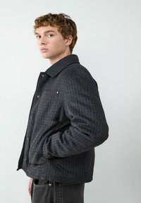 Grijze geruite jasje met een kraag, voorzakken en knoopsluitingen. Zachte textuur met een ontspannen pasvorm, gedragen over een zwart shirt.