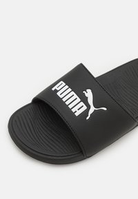 Sort slidesandal lavet af syntetisk materiale, med en bred rem med hvid "PUMA"-logo og tekstureret sål for godt greb.