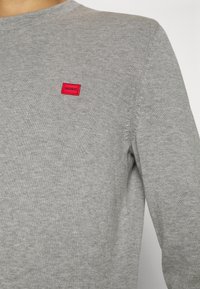 Gros plan d'une personne portant un pull en maille gris clair avec une petite étiquette rectangulaire rouge sur la poitrine indiquant "HUGO."