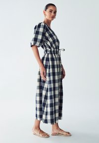 Vestido midi a cuadros en navy y crema con mangas abullonadas, cintura ceñida, aberturas laterales y un lazo. Combinado con sandalias minimalistas de tiras.