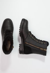 Botines negros hasta el tobillo con un upper de cuero suave, punta redonda y detalles de ante negro texturado. Presentan ojales dorados, una cremallera en el lateral y suela resistente.
