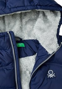 Chaqueta acolchada de color azul marino con un forro interior gris y esponjoso. Presenta una cremallera plateada y un logotipo bordado en la parte frontal.