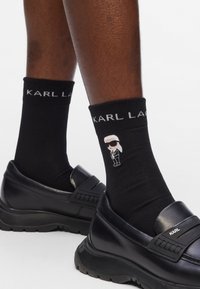 Calzini neri con il testo bianco "KARL LAGERFELD" e una figura cartone. Indossati con scarpe nere slip-on con una suola spessa e testurizzata.