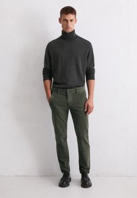 Dunkelgrauer Rollkragenpullover, enganliegend. Olivgrüne Cordhosen, schmal geschnitten. Schwarze Schnürschuhe, glatte Textur. Einfacher Hintergrund.