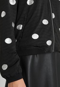 Giacca bomber nera con texture, caratterizzata da pois argentati, chiusura con zip e polsini elasticizzati. Accompagnata da una gonna in pelle nera.