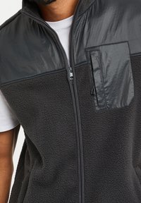 Fleece vest v tmavě šedé barvě s lesklým, hladkým yoke. Obsahuje zipové zapínání a kapsu na levé hrudi s elegantním designem.
