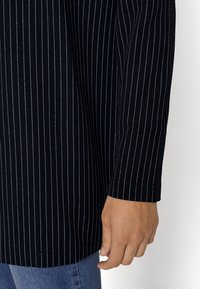 Czarna tkanina w białe paski, ukazująca mankiet długiego rękawa i dłoń go chwytającą. Gładka tekstura, dopasowany fason.