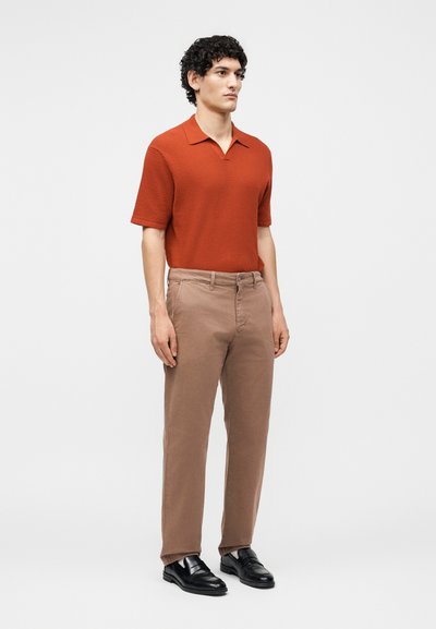 Homme debout portant un polo rouille à manches courtes texturé, un pantalon beige et des chaussures en cuir noir sans lacets sur fond blanc uni.