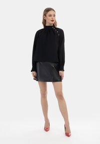 Schwarzes Langarmoberteil mit hohem Kragen und seitlichen Knopfdetails, kombiniert mit einem schwarzen Leder-Minirock und roten Lackpumps.